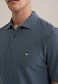 Polo paita slate blue värissä, jossa on teksturoitu kangas. Edessä vasemmalla rintapielessä musta-valkoinen brodeerattu lintulogo. Kaksi nappia napitusta.