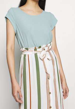 Femme portant un haut bleu clair à manches courtes rentré dans une jupe blanche taille haute avec des rayures verticales vertes et orange et une ceinture nouée.