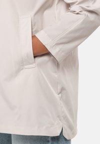 Helle beige Jacke aus glattem, wasserabweisendem Material. Verfügt über eine Seitentasche, hochgerollte Ärmel und einen geraden Saum.