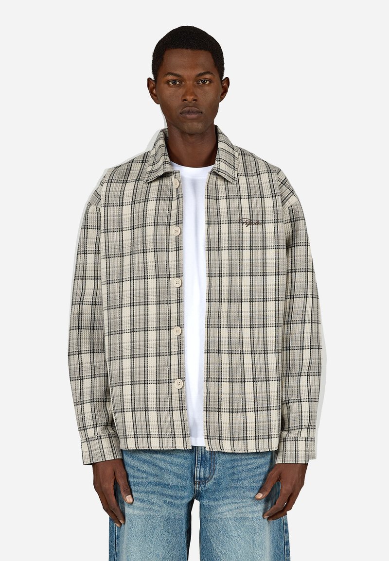 Beige és fekete kockás ing, gombos elülső rész, hegyes gallér, hosszú ujjak és mellkasi logó. Fehér póló és farmernadrág fölé van véve.