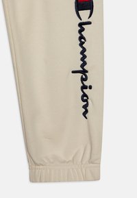 Beežid dressipüksid elastsete soonikutega, mille külgedel on silmatorkav must tikandiga "Champion" logo ja tekstuurne kangas.