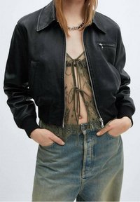 Femme portant une veste en cuir noire par-dessus un haut en dentelle vert olive transparent noué à l'avant, associée à un jean bleu taille haute, les mains dans les poches.