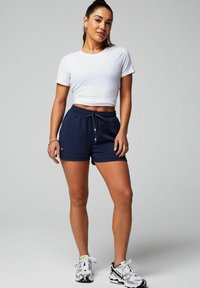 Weißes, kürzer geschnittenes T-Shirt kombiniert mit dunkelblauen Sportshorts, die über ein elastisches Bündchen und eine Kordel verfügen. Weiße und schwarze Sportschuhe.