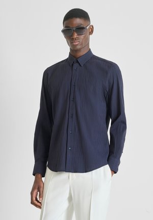 Uomo con camicia blu scuro a bottoni con trama, pantaloni bianchi e grandi occhiali da sole neri, in piedi contro uno sfondo chiaro e uniforme.