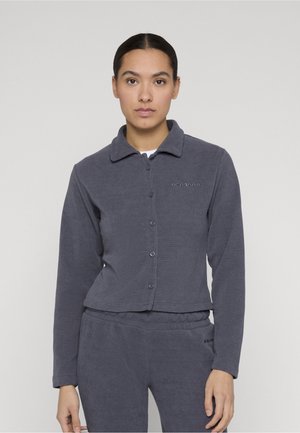 Donna con capelli scuri raccolti indossa una maglia a maniche lunghe grigio scuro con bottoni e pantaloni abbinati, in piedi davanti a uno sfondo chiaro uniforme.