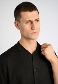 Svart stickad pikétröja med krage och knappslå. Tyget har en fin textur, med en diskret emblemmärkning på vänster sida.