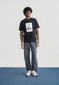 Μαύρο βαμβακερό t-shirt με γραφικό σχέδιο από αθλητικά παπούτσια. Συνδυάζεται με γκρι παντελόνι με φαρδιά μπατζάκια και λευκά αθλητικά παπούτσια με μαύρες λεπτομέρειες.