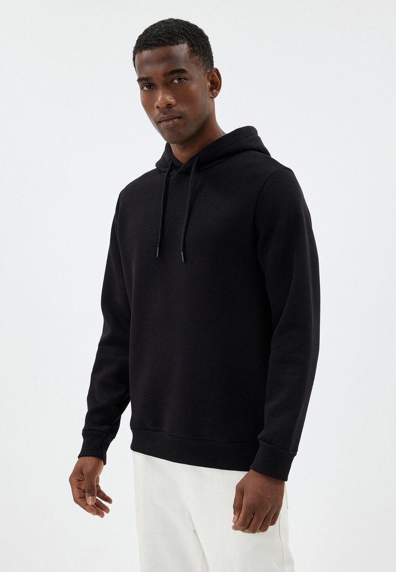 Sudadera negra con capucha ajustable y puños de canalé, hecha de una tela suave. El diseño incluye un bolsillo tipo canguro y un corte relajado.