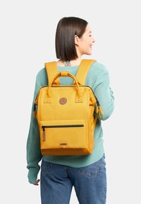 Zaino in tela giallo con tasca anteriore con zip, doppi manici in alto, spallacci imbottiti e cinghie laterali per accessori. Finitura testurizzata.