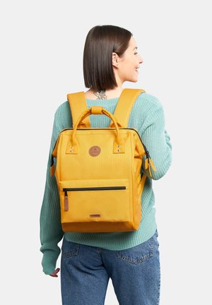 Zaino in tela giallo con tasca anteriore con zip, doppi manici in alto, spallacci imbottiti e cinghie laterali per accessori. Finitura testurizzata.
