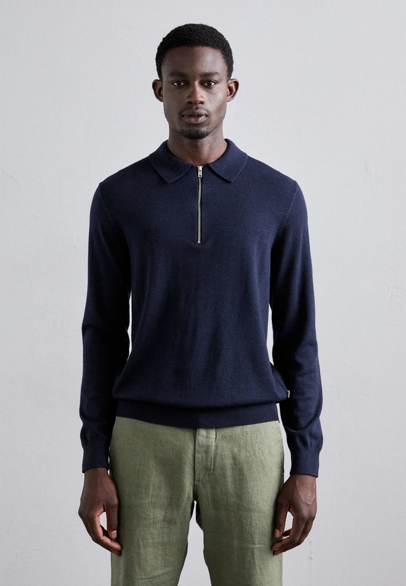 Marineblaues Langarm-Polohemd mit einem Viertel-Reißverschluss-Kragen, gerippten Bündchen und Saum; kombiniert mit hellgrünen Leinenhosen. Sanfte Textur.