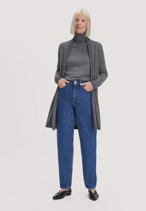 Frau mit glattem blondem Haar, die einen grauen Rollkragenpullover und eine Strickjacke, blaue Jeans und schwarze Schuhe trägt und mit den Händen in den Taschen steht.