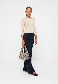 Cárdigan de punto beige abotonado en la parte frontal, combinado con pantalones de pierna ancha oscuros. Lleva un bolso de estampado de piel de serpiente. Zapatos oscuros completan el look.