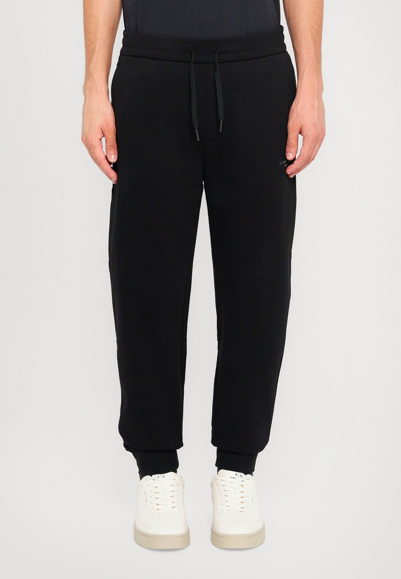 Sorte joggebukser med elastisk midjebånd, snor, sidelommer og ribbede mansjetter. Kombinert med hvite sneakers for kontrast.