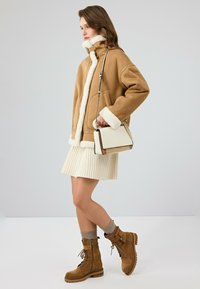 Bruine shearling vliegtuigjack met witte rand, ivoorkleurige geplooide rok en bruine veterschoenen, accessorized met een kleine crèmekleurige handtas.