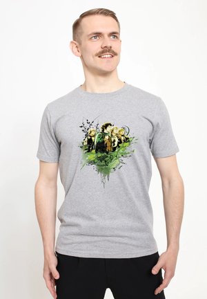 Uomo con baffi che indossa una maglietta grigia con quattro personaggi illustrati circondati da fogliame verde ed elementi astratti neri.