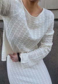 Valge kudum, millel on lainemuster, pikad varrukad ja ümmargune kaelus, koos sarnase tekstuuriga seelikuga.