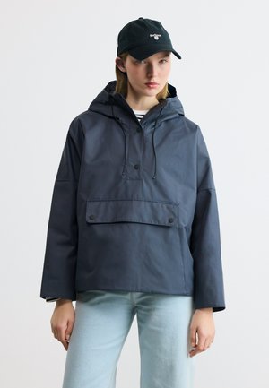 Frau trägt eine marineblaue Anorakjacke mit Fronttasche und Kapuze, hellblaue Jeans und eine schwarze Barbour-Baseballkappe.