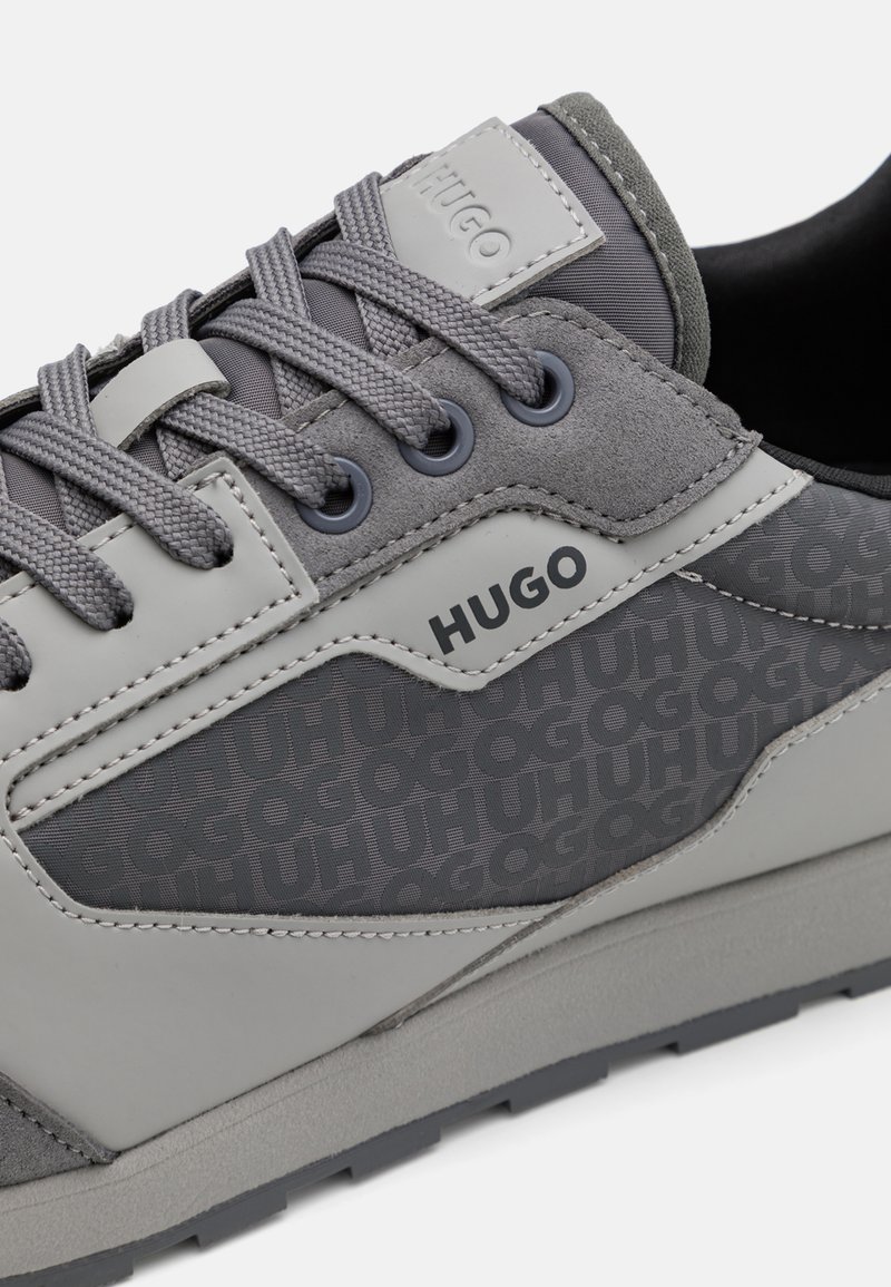 Graue Sneakers aus einer Mischung aus matten und strukturierten Materialien, mit dezentem Branding, kontrastierenden Überlagerungen und einem gemusterten Seitenpaneel.