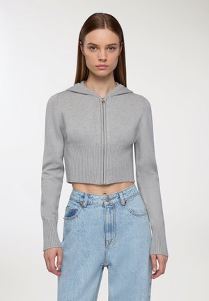 Felpa grigia chiara cropped con orlo a coste e maniche lunghe, caratterizzata da una zip frontale e una texture morbida, abbinata a jeans blu a vita alta.