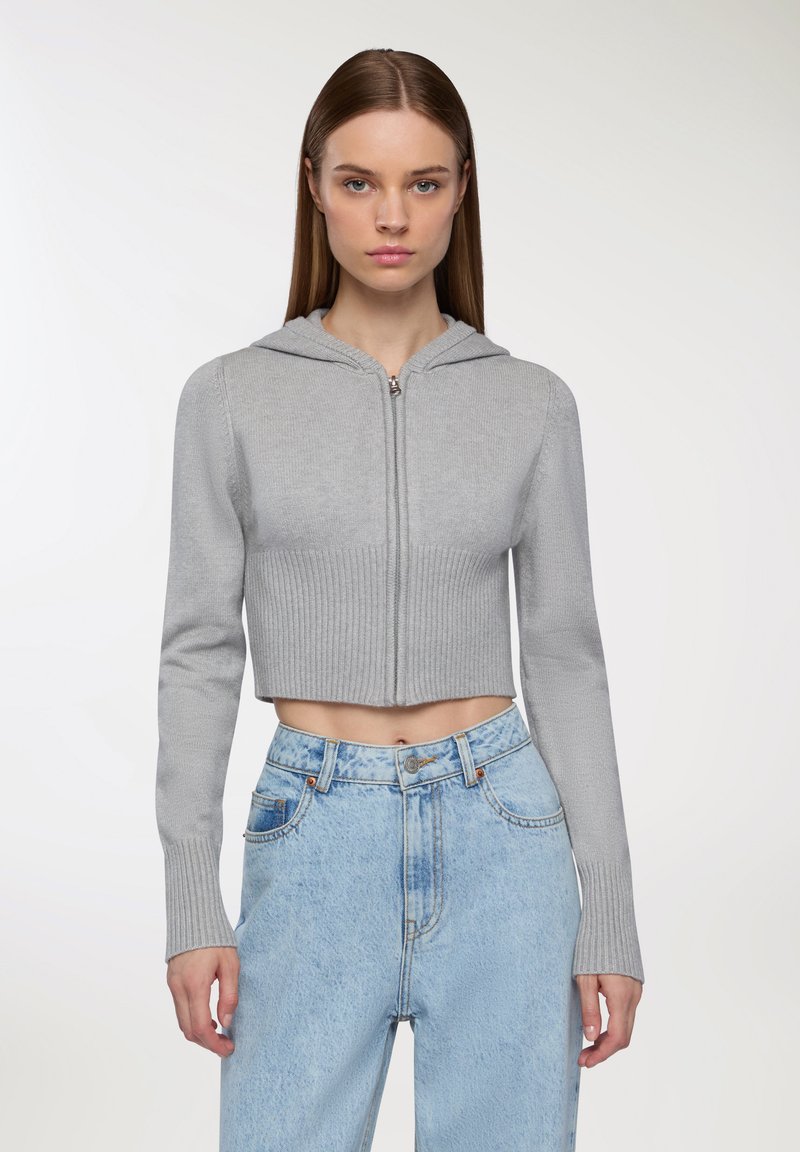 Felpa grigia chiara cropped con orlo a coste e maniche lunghe, caratterizzata da una zip frontale e una texture morbida, abbinata a jeans blu a vita alta.