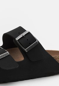 Sandales Birkenstock noires avec deux sangles réglables, boucles métalliques et une semelle en liège. Matériau doux et texturé, forme contourée.