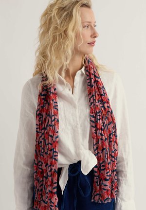 Blondínka v bielej košeli na gombíky zviazanej v páse, v námorníckych nohaviciach a s dlhým červeno-námorníckym kvetinovým šálom, pozerajúca sa do strany.