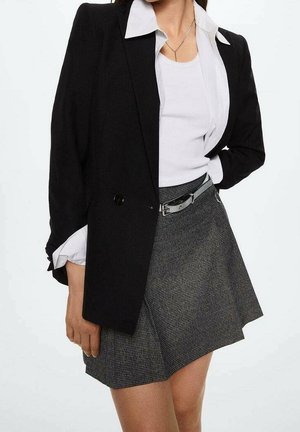 Woman wearing a black blazer over a white shirt and tank top, paired with a gray pleated skirt and a silver belt.