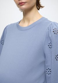 Svijetloplavi pulover s okruglim izrezom i napuhanim rukavima, ukrašen cvjetnim izrezanim uzorcima na rukavu. Mekana tekstura tkanine.