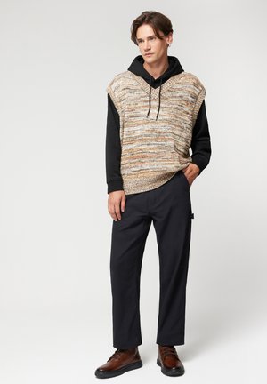 Gruba, pleciona kamizelka z wielokolorowej przędzy na czarnej bluzie z kapturem, w połączeniu z ciemnymi spodniami i brązowymi sznurowanymi butami.