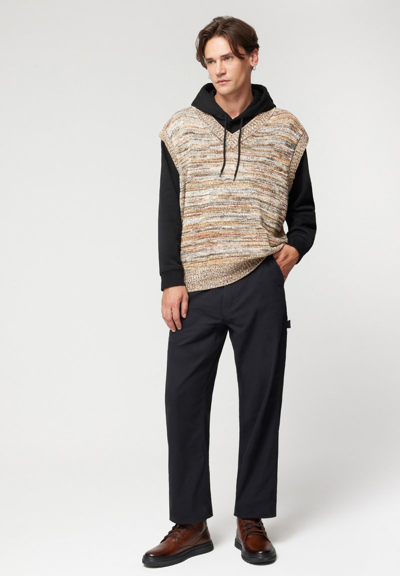 Gruba, pleciona kamizelka z wielokolorowej przędzy na czarnej bluzie z kapturem, w połączeniu z ciemnymi spodniami i brązowymi sznurowanymi butami.