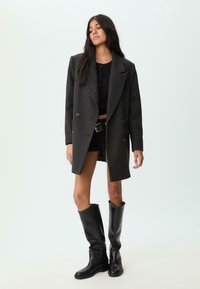 Dobbeltknappet grå uldfrakke med store revers, båret over en sort cropped top og shorts. Sammen med støvler i sort læder op til knæene.
