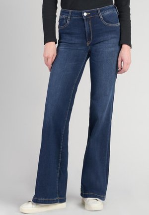 Jean évasé bleu foncé en denim. Présente des surpiqûres marquées, cinq poches et une fermeture à bouton à l'avant.