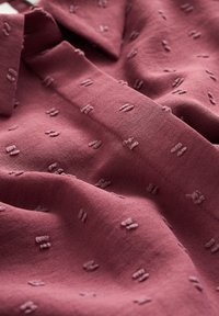 Burgundin kangas, jossa on teksturoitu kuvio, sisältää pieniä koholla olevia brodeerattuja muotoja, pehmeä pinta ja siisti kaulustyyli.