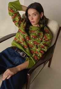Knit green sweater with orange fox pattern, ribbed cuffs, worn with blue high-waisted jeans and a brown belt, seated in a wooden chair.