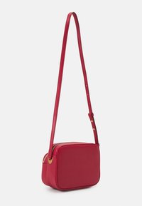 Červená kůže křížová taška s obdélníkovým tvarem, texturovaným povrchem, štíhlým nastavitelným popruhem a zlatými akcenty z kovu.
