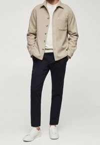 Veste chemise en mélange de laine beige avec des boutons, poche poitrine unique, associée à un pantalon bleu marine et des baskets blanches. Design simple et moderne.