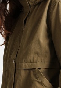 Olivgrön, lätt jacka med huva, som har knappstängning och sidofickor. Tyget har en slät textur och matt finish.