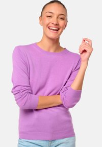 Femme souriante portant un pull lilas à manches longues et un jean bleu clair, avec un bras croisé et l'autre main levée.