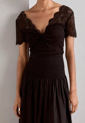 Vrouw draagt een zwarte jurk met kanten korte mouwen, een gedraaide halslijn aan de voorkant en een taille met smockwerk, met een vloeiende rok.