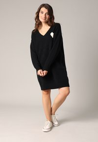 Robe noire en tricot avec un col en V et des manches longues, arborant un accent en forme de cœur blanc sur le côté gauche de la poitrine. Associée à des baskets blanches.