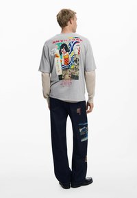 Šedé tričko s farebnou grafikou na zadnej strane, v kombinácii s tmavými džínsami so záplatami a voľným strihom.