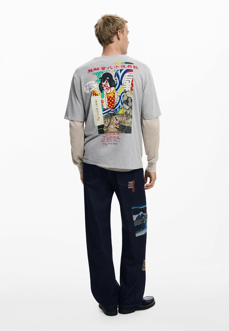Šedé tričko s farebnou grafikou na zadnej strane, v kombinácii s tmavými džínsami so záplatami a voľným strihom.