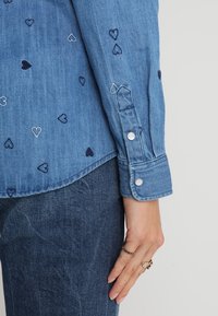 Spijkershirt met korte opgerolde mouwen, voorzien van geborduurde, meerkleurige harten. Lichtblauwe stof met een gladde textuur en zilveren drukknopen.