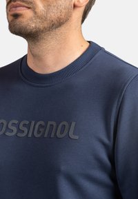Sudadera azul marino con cuello redondo acanalado, textura suave y un logo negro prominente en el pecho.