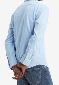 Chemise bleu clair à manches longues en tissu doux, avec poignets boutonnés et col classique, associée à un jean bleu foncé avec des poches arrière.