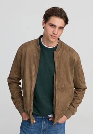 Giacca bomber in suede marrone con colletto a costine, zip frontale e una texture morbida, indossata sopra un maglione in maglia verde scuro.