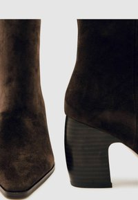 Brūni zamšādas potītes zābaki ar izteiktu degunu, ar masīvu svītrotu melnu papēdi un gludu tekstūru.