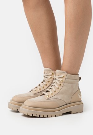 Botas de tornozelo em camurça bege com sola de borracha grossa, ilhoses dourados e design de atacadores. Apresenta um colarinho acolchoado e um detalhe de logótipo na lateral.