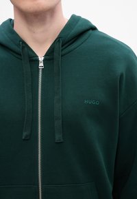 Grøn zip-up hoodie lavet af bomuld, med en struktureret hætte, to snore og et lille "HUGO" logo på brystet.